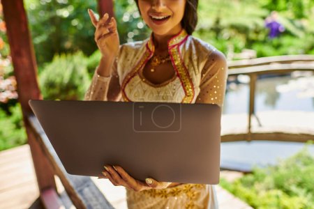 abgeschnittene Ansicht einer indischen Frau im authentischen Stil, die während eines Videogesprächs am Laptop an einem Sommertag spricht