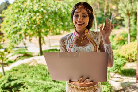 Foto de Mujer india alegre en traje tradicional saludando la mano durante la videollamada en el portátil en el parque de verano - Imagen libre de derechos