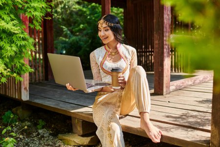 glückliche indische Frau in ethnischer Kleidung mit Laptop und Kaffee zum Mitnehmen in einer hölzernen Nische im Park