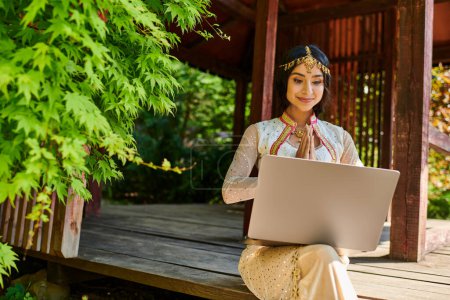 smiling indian woman in traditional clothes showing greeting gesture during video call on laptop