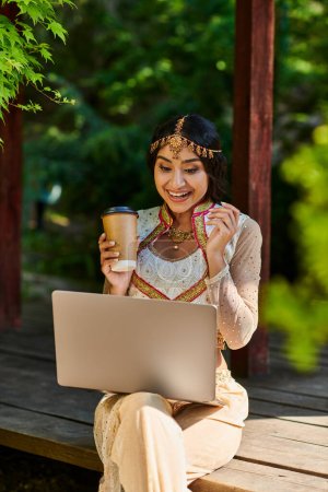fröhliche indische Frau mit Coffee to go und Blick auf Laptop in hölzerner Nische im Park an einem Sommertag
