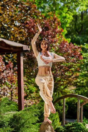brunette indian woman in authentic style attire dancing on park on sunny summer day