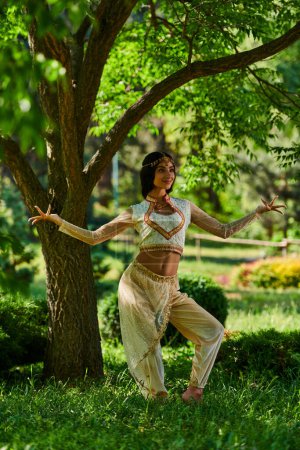 anmutige indische Frau in lebendiger ethnischer Kleidung tanzt unter einem Baum im Sommerpark, Glück