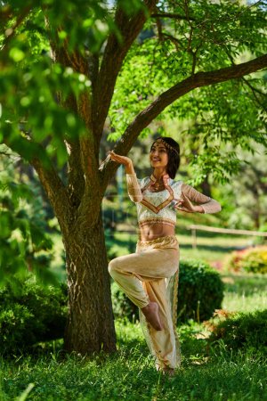 happy indian woman in elegant authentic attire dancing under tree on green lawn, summer sunny park