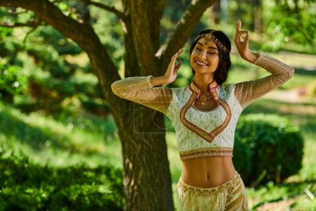 junge und glückliche indische Frau in authentischer Kleidung tanzt im Park an einem sonnigen Tag, Sommerglück
