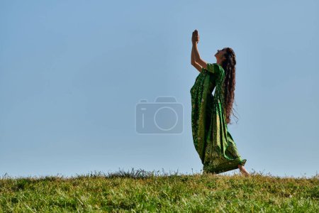 Téléchargez les photos : Joie d'été, femme indienne en vêtements authentiques, avec les mains priantes sur la pelouse verte sous le ciel bleu - en image libre de droit