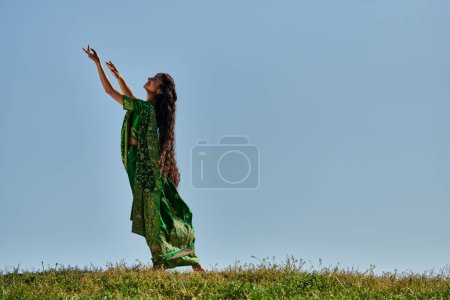 Sommertag, indische Frau in authentischer Kleidung mit ausgestreckten Händen im grünen Feld unter blauem Himmel