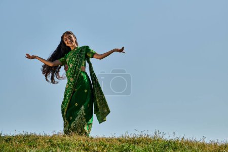 Unbekümmerte Indianerin im traditionellen Sari lächelt auf grünem Rasen unter blauem Himmel in die Kamera