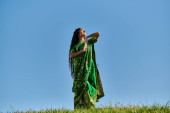 summer enjoyment, green field, indian woman in ethnic wear smiling with closed eyes under blue sky hoodie #671993786