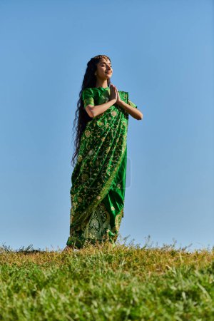 happy indian woman in sari with praying hands and closed eyes on lawn under blue sky, summer day