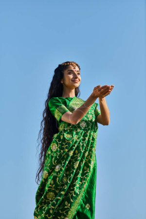 verano, día soleado, mujer asiática alegre con las manos extendidas de pie en sari bajo el cielo azul