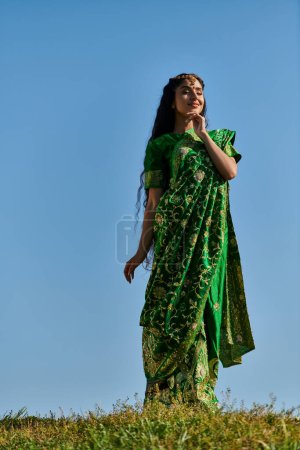 junge indische Frau im eleganten traditionellen Sari im grünen Feld unter blauem Himmel, Sommerglück