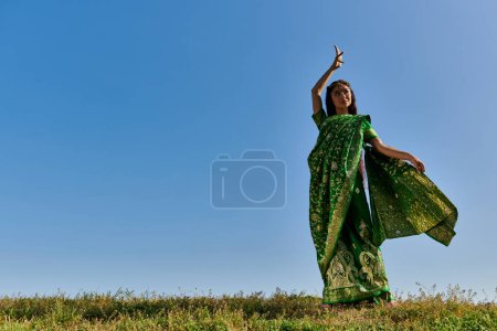 summer dance of smiling indian woman in authentic sari in green field under blue sky