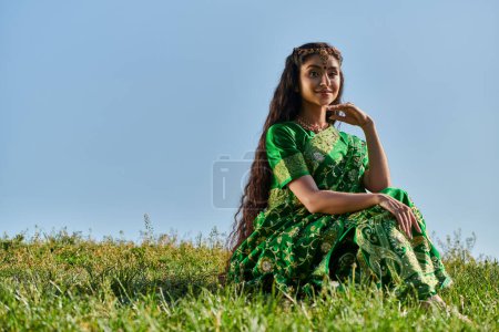 mujer india en ropa étnica, sari, sentado en el césped verde bajo el cielo azul del verano y sonriendo a la cámara
