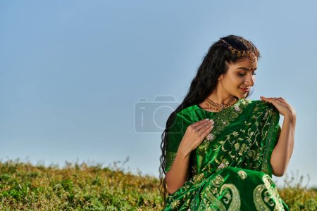 young indian woman with matha patti touching green sari on hill with blue sky at background
