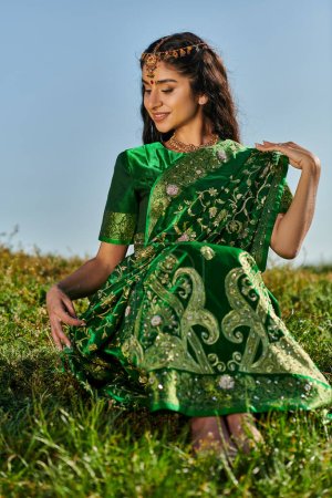 smiling young indian woman touching green sari while sitting on grassy hill with sky on background