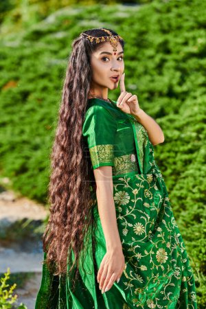 junge Indianerin im Sari zeigt geheime Geste und blickt in die Kamera in der Nähe von Pflanzen im Park