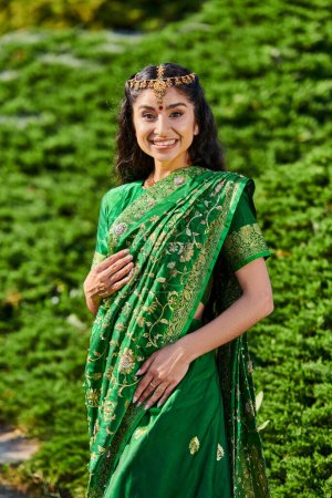 Porträt einer jungen fröhlichen indischen Frau in traditionellem Sari und matha patti, die im Park in die Kamera blickt