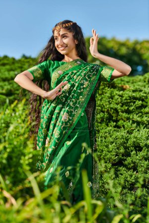 carefree young indian woman in traditional sari dancing while standing near plants in park