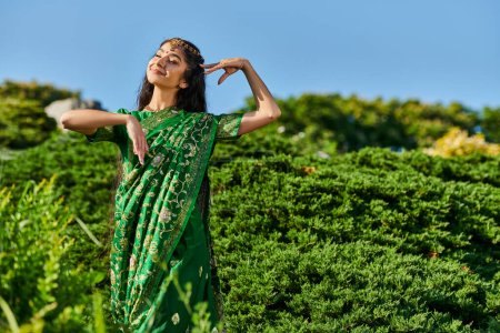 pretty young indian woman in sari and matha patti posing near plants in park on background