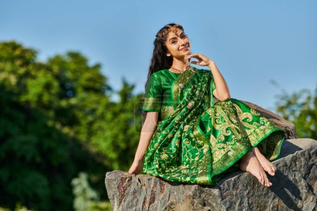 smiling and stylish indian woman in sari posing while sitting on stone with blue sky on background