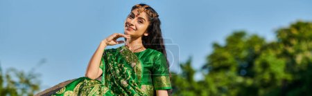 smiling indian woman in green sari and matha patti posing at camera with sky on background, banner