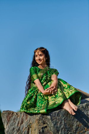 Foto de Despreocupado joven india mujer en verde sari sentado en piedra y mirando hacia otro lado con el cielo en el fondo - Imagen libre de derechos