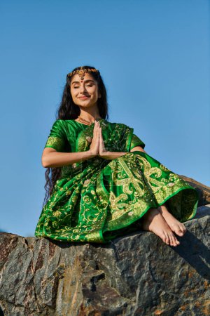 smiling indian woman in sari doing praying hands gesture on stone with blue sky on background