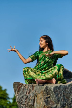 fröhliche und stilvolle indische Frau in grünem Sari posiert auf Stein mit blauem Himmel auf Hintergrund
