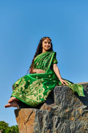 carefree barefoot indian woman in stylish green sari sitting on stone with sky on background