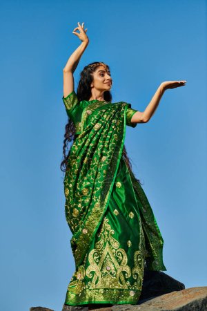 vue à angle bas de femme indienne positive en sari posant sur la pierre avec ciel bleu à l'arrière-plan