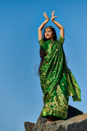 cheerful young indian woman in green sari posing on stone with blue sky on background