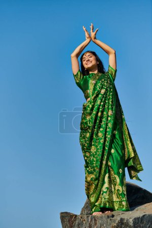 low angle view of smiling indian woman in sari posing while standing on stone with sky on background