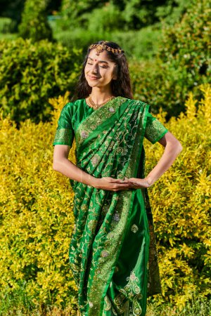 Foto de Mujer india joven elegante y sonriente en sari posando en arbustos en el parque al aire libre - Imagen libre de derechos