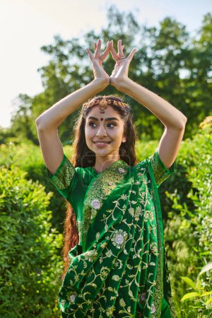 fröhliche junge Indianerin in grünem Sari und Bindi gestikulierend neben Pflanzen im Park
