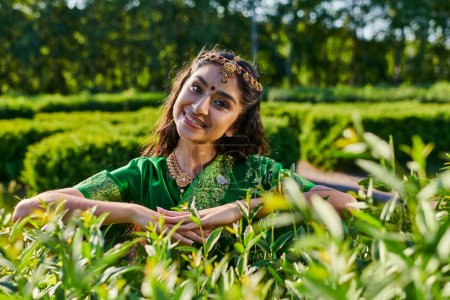 pretty young asian woman in stylish sari and bindi looking at camera near plants in park