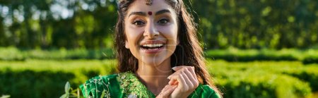 pretty young indian woman in sari smiling at camera near blurred plants outdoors, banner