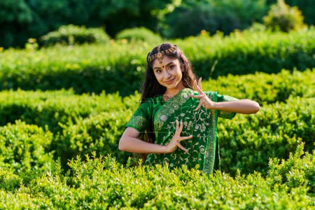 mujer india joven de moda en sari verde sonriendo y posando cerca de arbustos en el parque en verano
