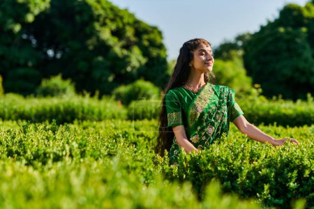 erfreut junge Indianerin im Sari, die mit geschlossenen Augen neben grünen Pflanzen im Park steht