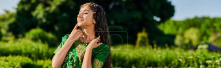 joyful young indian woman in matha patti and sari standing near blurred plants in park, banner