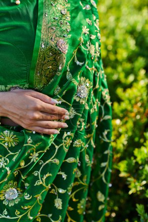 cropped view of young woman in stylish sari with pattern standing near green bushes outdoors