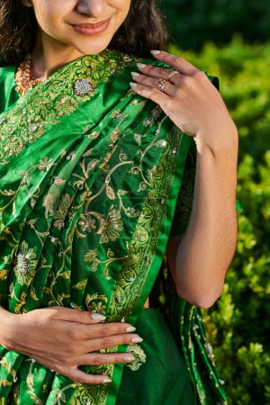 Teilbild einer lächelnden und eleganten jungen Frau im modernen Sari, die neben verschwommenen Pflanzen im Freien steht