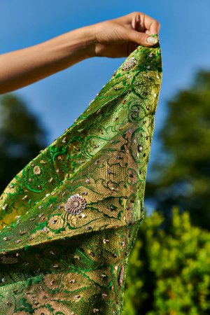 vista recortada de mujer joven sosteniendo sari verde moderno con patrón cerca de plantas borrosas al aire libre