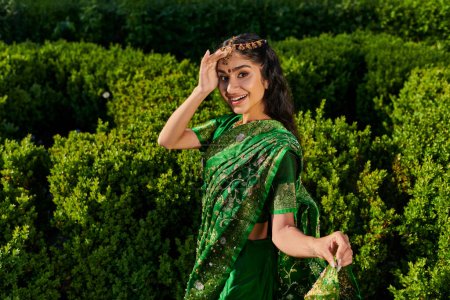 excited and stylish indian woman in sari with pattern and matha patti near green plants outdoors