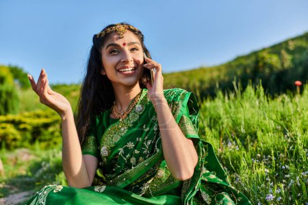 smiling young indian woman in green sari talking on smartphone while sitting on grass in summer
