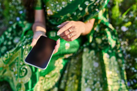 Ausgeschnittene Ansicht einer jungen Frau im grünen Sari, die im Sommer ihr Smartphone auf dem Rasen hält