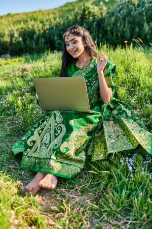 mujer india con estilo en sari tener videollamada en el ordenador portátil mientras está sentado en el prado en verano
