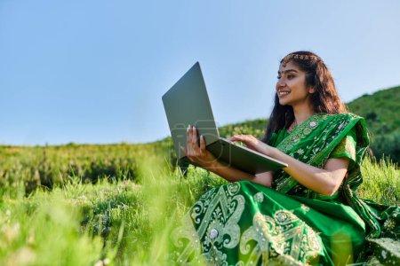 sonriente joven india mujer en sari verde usando portátil en prado herboso en verano