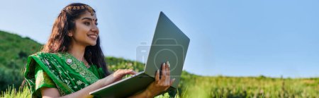 stylish young indian woman in green sari using laptop on grassy field in summer, banner