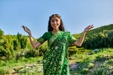 cheerful young indian woman in green sari posing and looking at camera on summer field